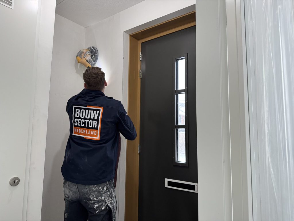 Vakman in een donkerblauwe jas van Bouwsector Nederland schuurt de wand boven een voordeur glad