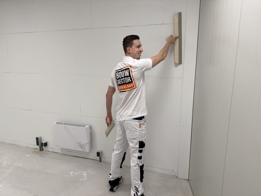 Lachende stukadoor van Bouwsector Nederland met een lange spaan en een plakspaan aan een grote wand
