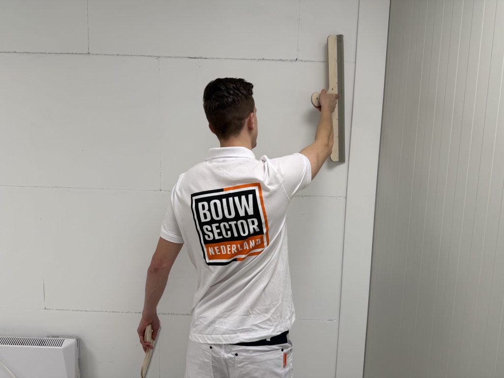 Stukadoor in wit werkshirt met logo van Bouwsector Nederland zet een wand vlak met een lange spaan