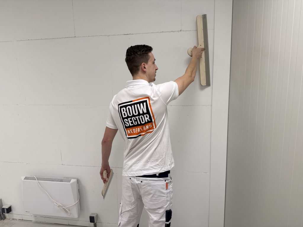 Stukadoor trekt met een lange spaan een wand vlak in een lege kamer met een radiator