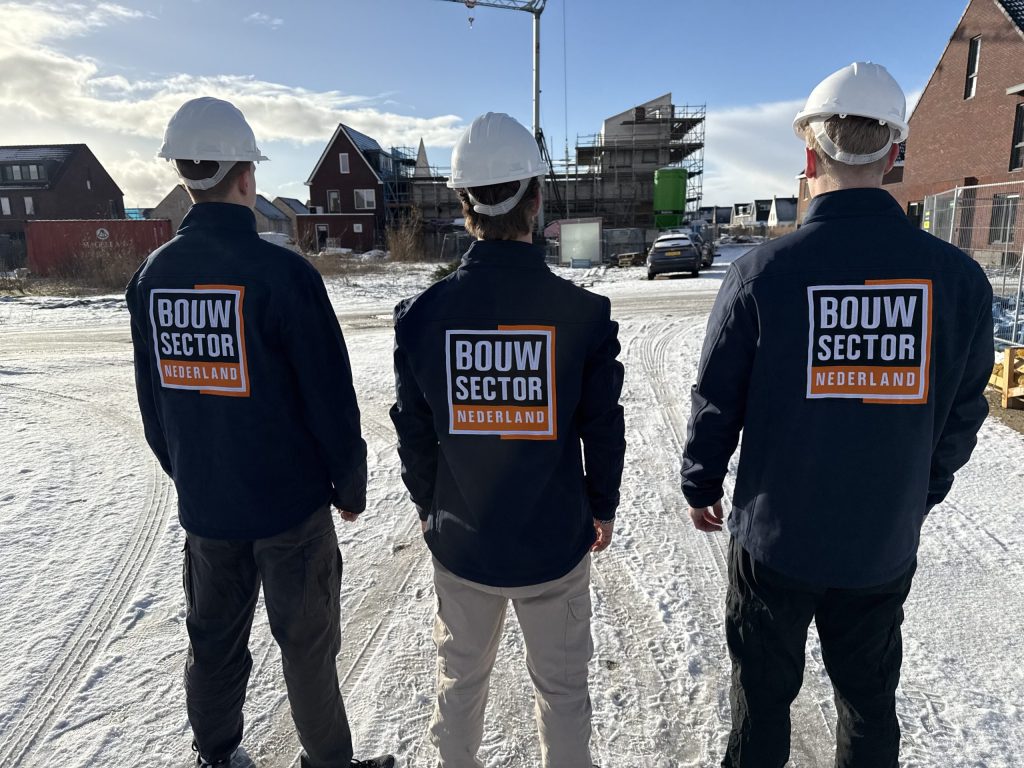 Drie vakmensen van Bouwsector Nederland met witte helm kijken naar woningen in aanbouw op een besneeuwd terrein